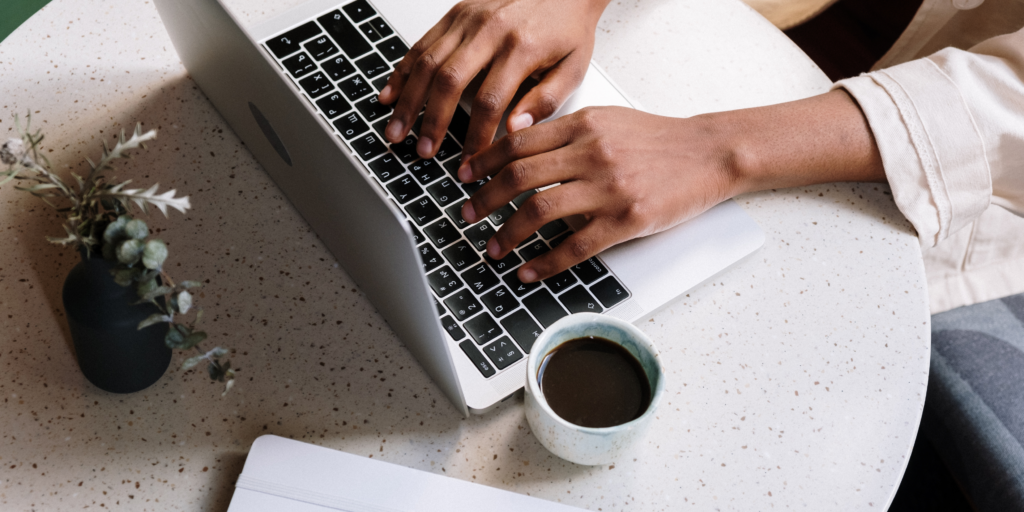 Close-up of a person typing on a MacBook with a cup of coffee on a round white table — ideal image for troubleshooting MacBook trackpad issues.
