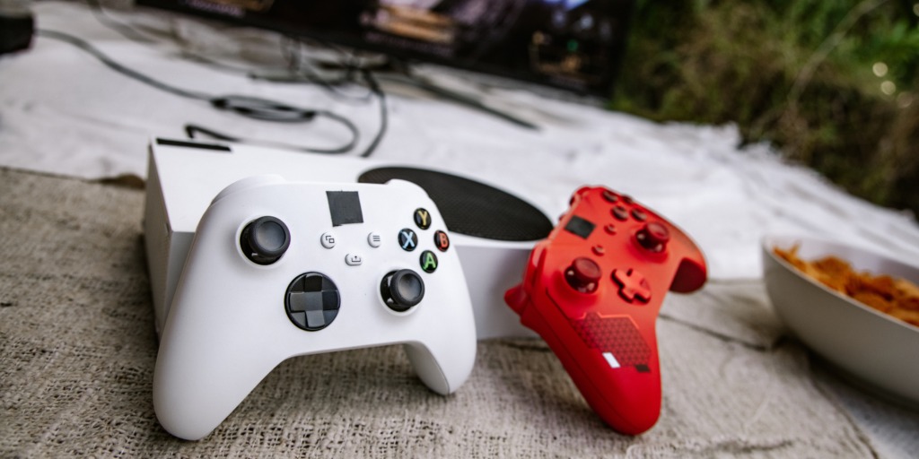 White and red Xbox controllers next to a console on a fabric surface — representing gaming console setup and crash-related issues. 
